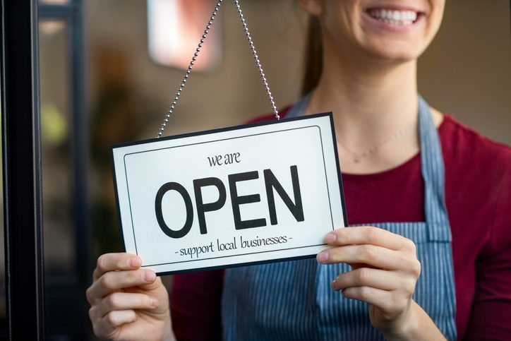 Open sign in a small business shop Small business owner smiling while turning the sign for the reopening of the place after the quarantine due to covid-19. Happy businesswoman standing at her restaurant or coffee shop gate with open signboard. Close up of woman"u2019s hands holding sign now we are open support local business.